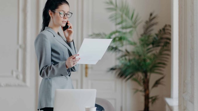 Serious brunette female employee stands near desktop, holds paper documents, talks via smartphone, wears formal clothes and spectacles, discusses business strategy with partner, organizes meeting